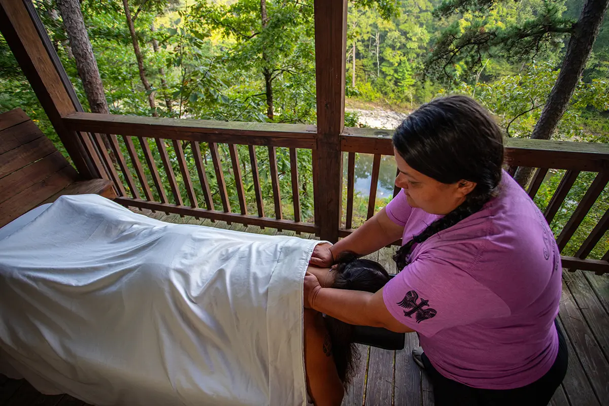 A massage therapist gives a neck massage on an outdoor wooden deck overlooking a green forest.